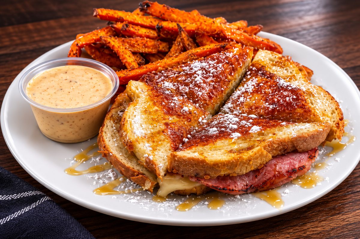 Grilled ham-and-cheese style sandwich dusted with powdered sugar and drizzled with syrup, with sweet potato fries and fry sauce